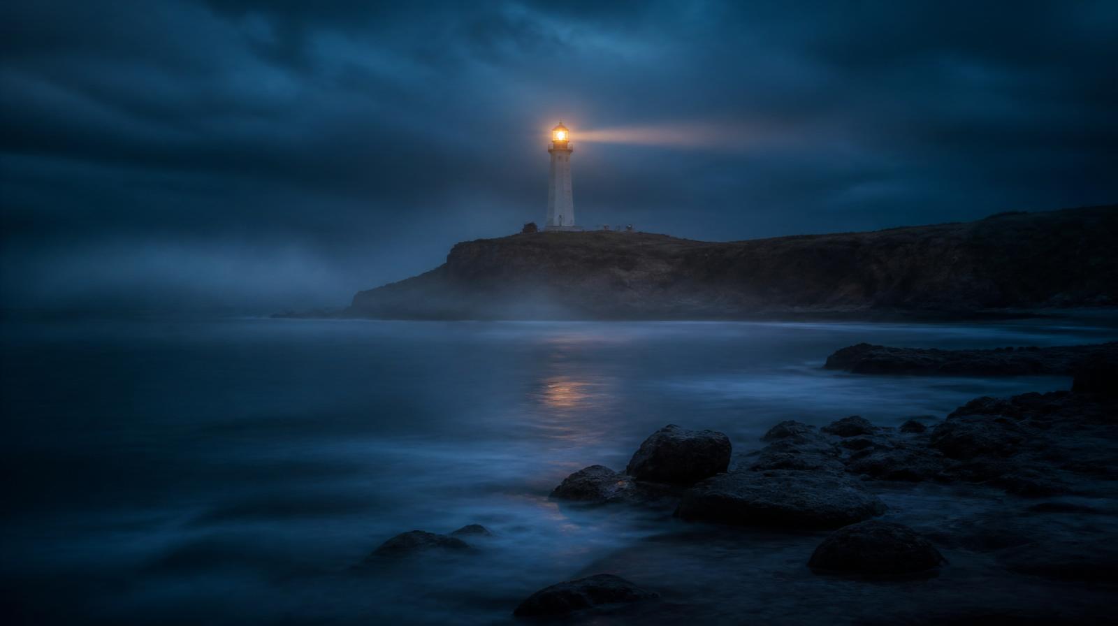 A lighthouse on a South African coastline at blue hour, beam cutting through mist.